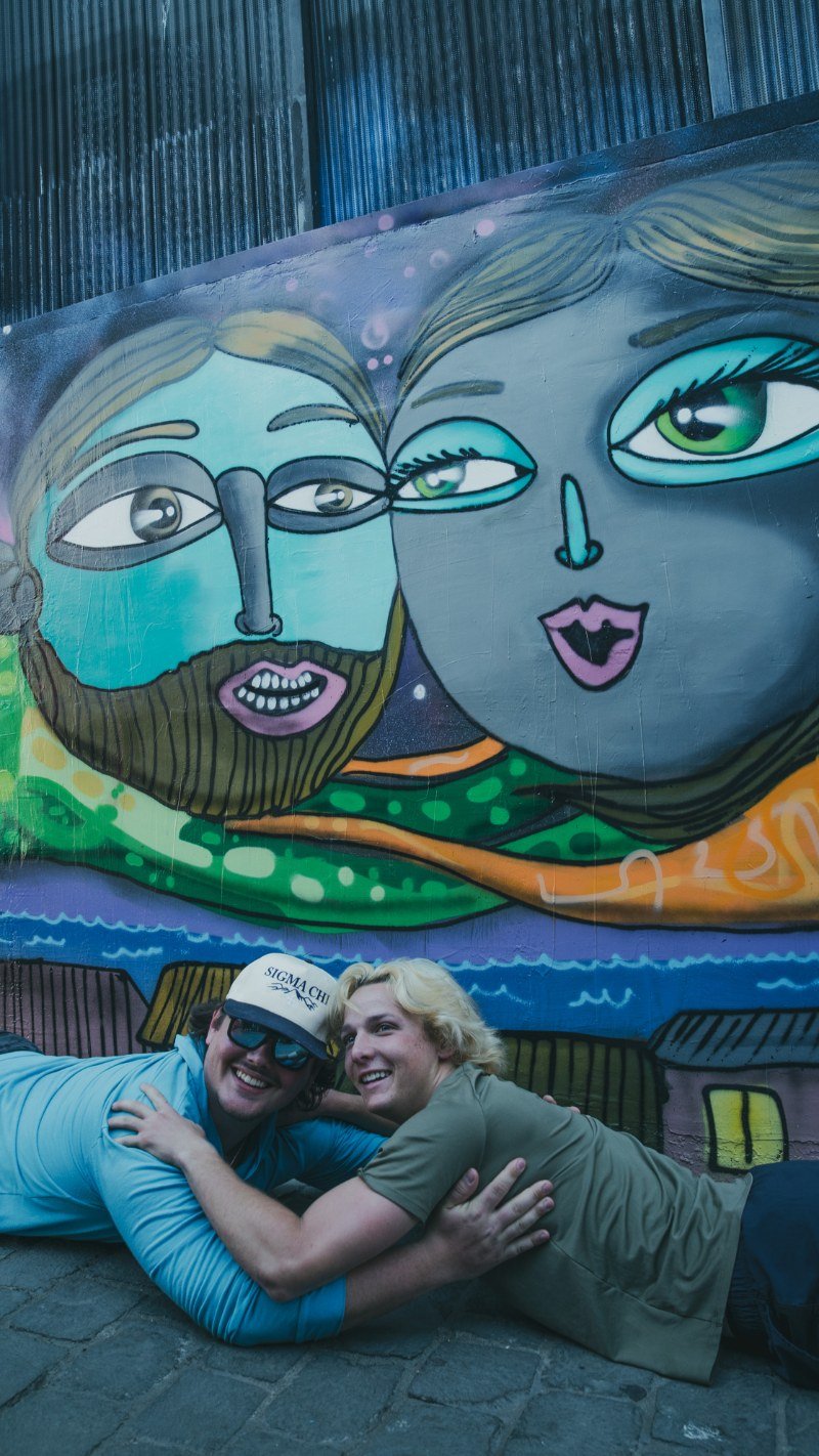 Two travelers posing under a colorful street-art mural of two faces in Valparaíso