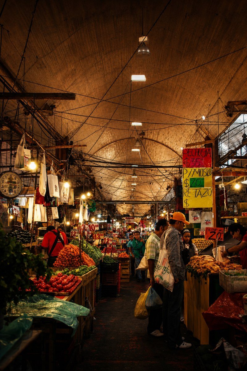 Shoppers walking the produce aisles of a covered market hall, stalls piled with fruit and vegetables