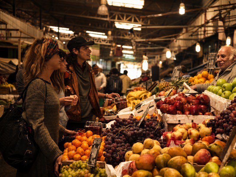 Travelers tasting fruit with a vendor at a produce stall in La Vega market, Santiago